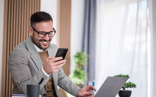 Mann sitzt am Schreibtisch und schaut auf sein Smartphone. Vor ihm steht sein Laptop