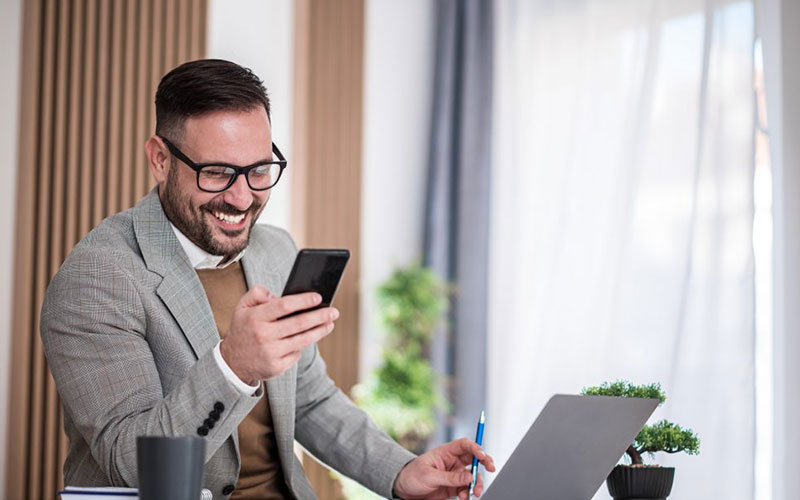 Mann sitzt am Schreibtisch und schaut auf sein Smartphone. Vor ihm steht sein Laptop