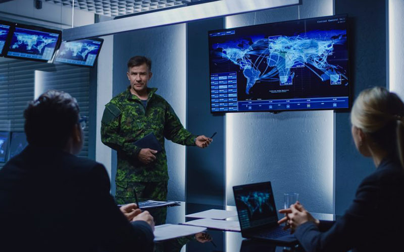 An officer in camouflage uniform presents a digital world map with networks and connections on a large screen in a modern meeting room. Two people in suits listen attentively while laptops and monitors display additional data